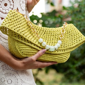 May include: A light green crocheted clutch with a gold chain strap and a beaded accent. The bag has a folded flap and is held by a person wearing a white crocheted dress. The background is blurred green foliage.