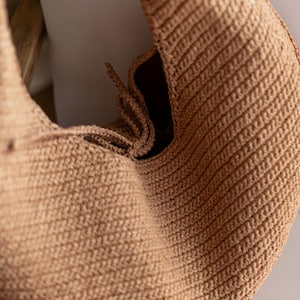 May include: A tan-colored, crocheted bag with a rounded shape. The bag features a textured, ribbed pattern and a drawstring closure. The interior is dark, contrasting with the lighter exterior. The bag is likely handmade.