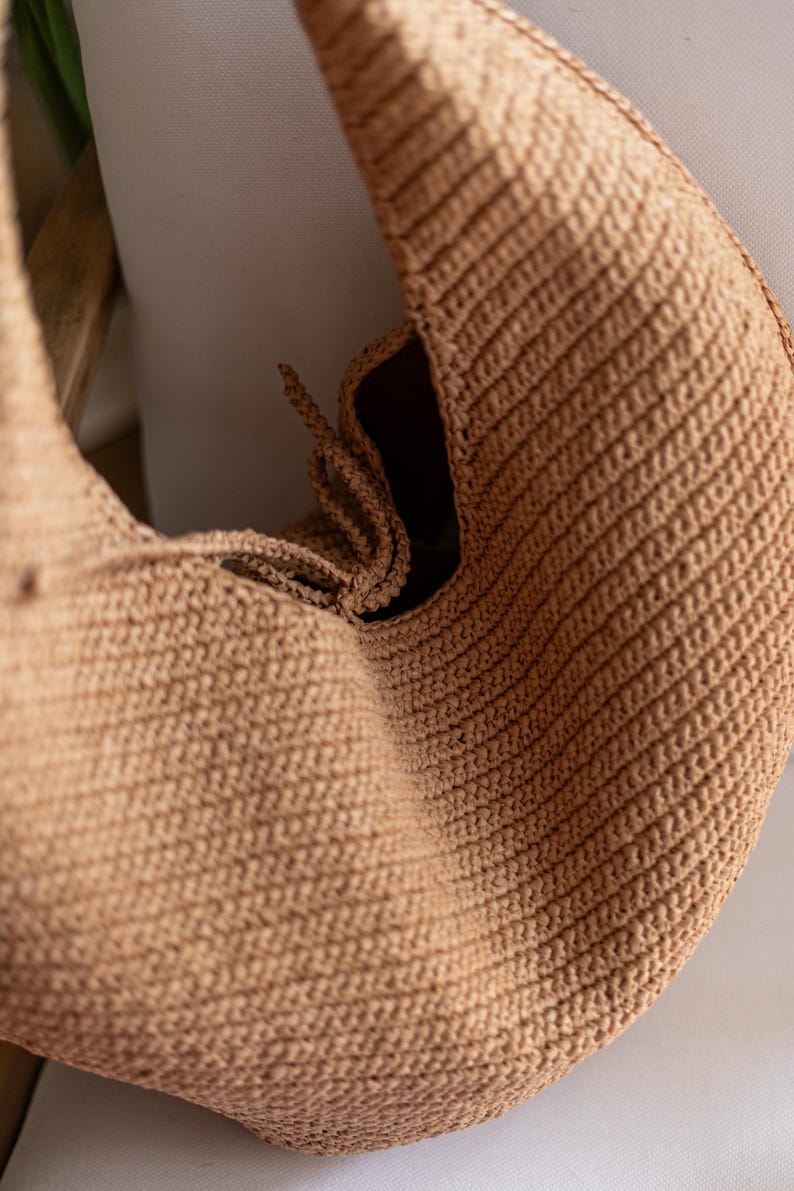 May include: A tan-colored, crocheted bag with a rounded shape. The bag features a textured, ribbed pattern and a drawstring closure. The interior is dark, contrasting with the lighter exterior. The bag is likely handmade.