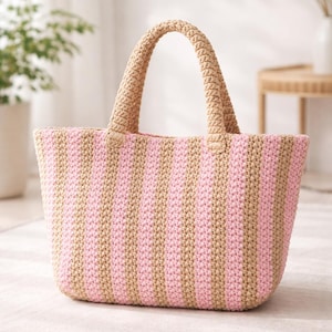 May include: A handmade tote bag with a striped pattern in pink and tan. The bag features two tan handles and is made of a textured, woven material. The bag is designed for carrying items and has a spacious interior.