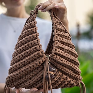 Puede incluir: Bolso marrón tejido a mano con forma triangular. El bolso presenta un asa trenzada y borlas decorativas. Un pequeño dije cuelga del asa. El bolso está hecho de un hilo grueso y texturizado.