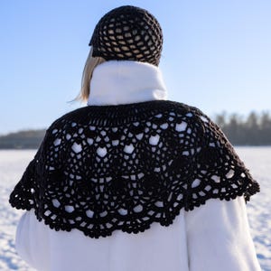 May include: A black crocheted capelet with an intricate openwork pattern featuring circular motifs and scalloped edges, worn over a white coat, paired with a matching black crocheted beanie with a grid-like texture on the head.