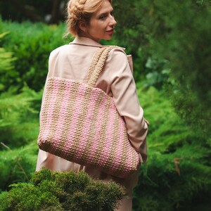 May include: A large, striped tote bag in shades of pink and tan, carried over the shoulder. The bag has a woven texture and a wide strap. The background features lush green foliage and a person wearing a tan coat.