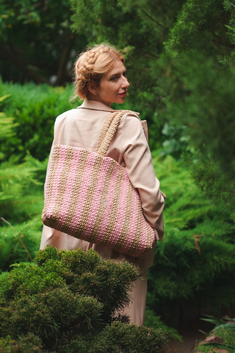 May include: A large, striped tote bag in shades of pink and tan, carried over the shoulder. The bag has a woven texture and a wide strap. The background features lush green foliage and a person wearing a tan coat.