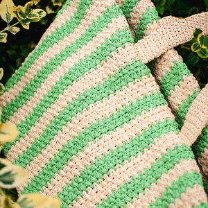 May include: A woven tote bag with alternating green and cream-colored horizontal stripes. The bag has matching cream-colored handles and is set against a backdrop of green foliage. The bag's texture is visible.
