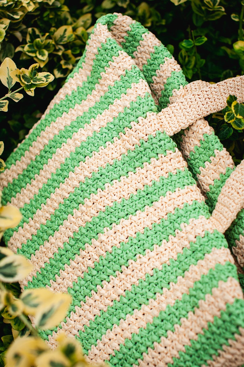 May include: A woven tote bag with alternating green and cream-colored horizontal stripes. The bag has matching cream-colored handles and is set against a backdrop of green foliage. The bag's texture is visible.