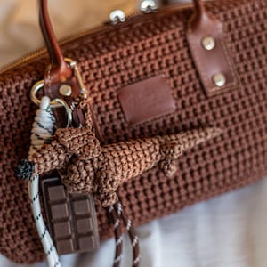 May include: A brown crochet handbag with a matching dachshund keychain and a chocolate bar charm. The bag has a woven texture, leather accents, and a zipper closure. The keychain is attached to a brown leather handle.