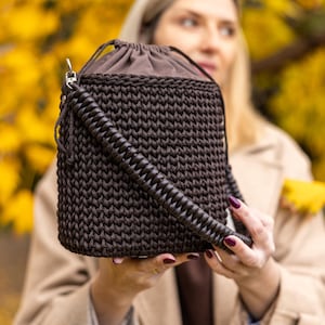 May include: A dark brown, hand-crocheted handbag with a drawstring closure and a thick, spiral-wrapped strap. The bag is a cube shape and appears to be made of a sturdy yarn. The background is blurred, with yellow foliage.