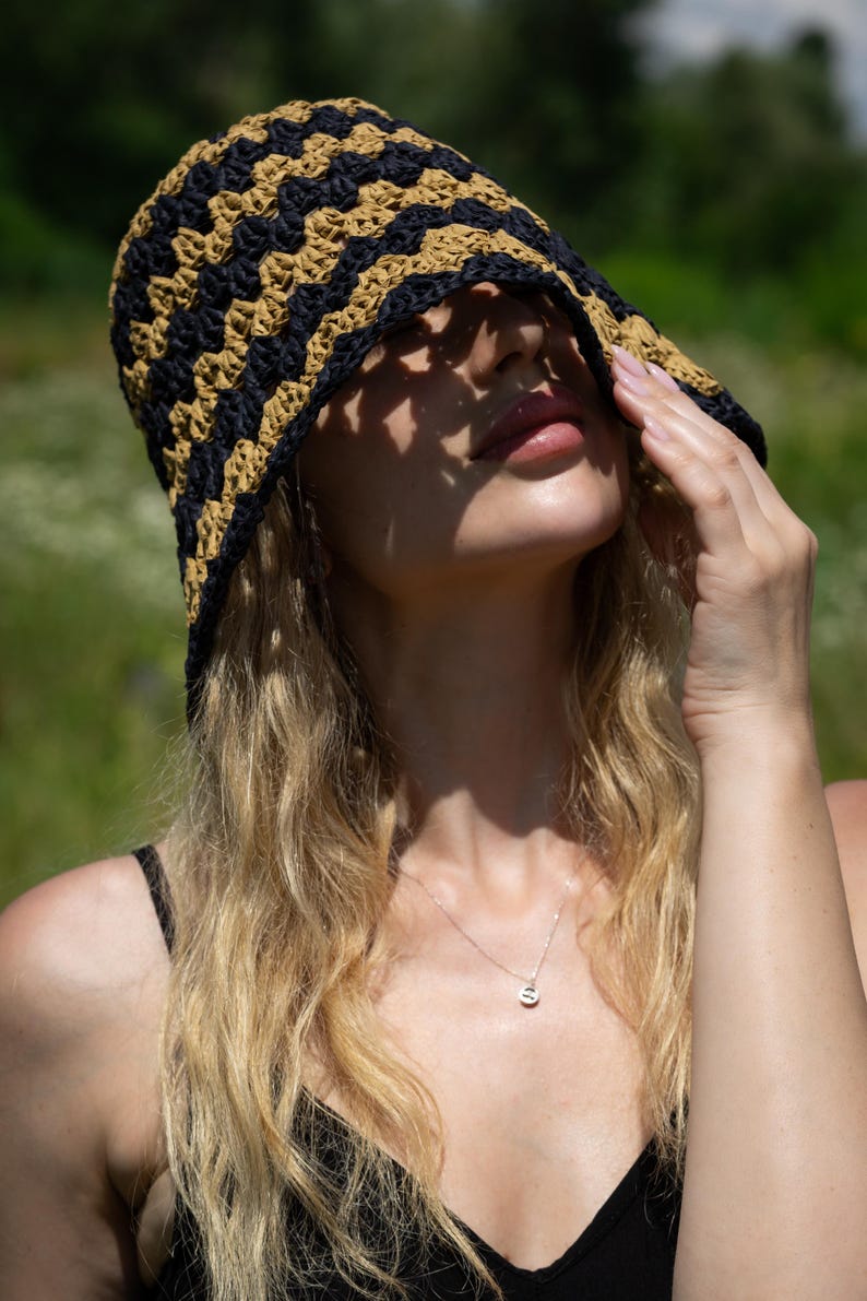 May include: A woman wearing a black and tan crocheted bucket hat with a brim that covers her eyes. She is wearing a black tank top and a silver necklace with a small pendant.