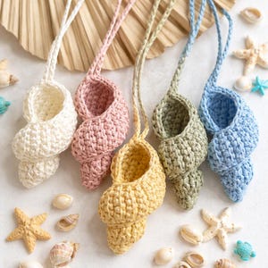 May include: Five crochet seashell ornaments in white, pink, yellow, green, and blue, each with a loop for hanging. The seashells are surrounded by starfish, seashells, and small turtle figurines, creating a beachy theme.