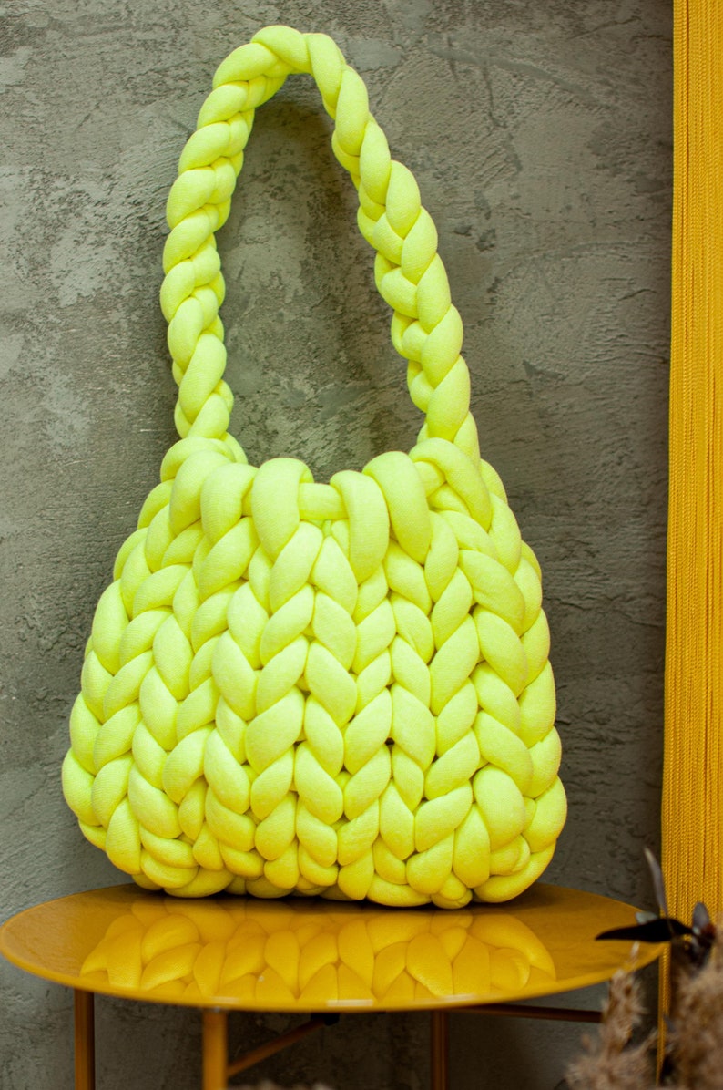 Puede incluir: Un bolso tote de punto amarillo brillante con asas trenzadas gruesas. El bolso est&aacute; hecho de un hilo suave y grueso y tiene un interior espacioso.