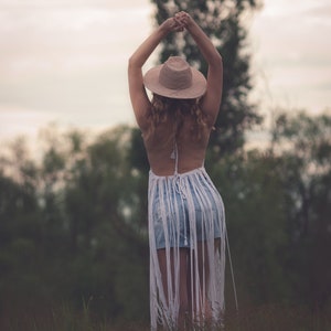 May include: A woman wearing a white crochet top with fringe, a straw hat, and denim shorts stands in a field of tall grass. She has her arms raised above her head.