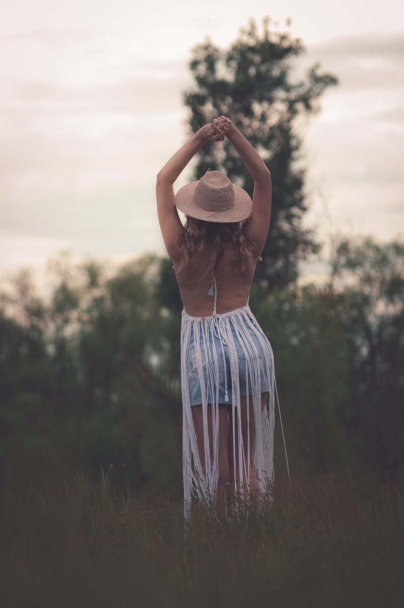 May include: A woman wearing a white crochet top with fringe, a straw hat, and denim shorts stands in a field of tall grass. She has her arms raised above her head.