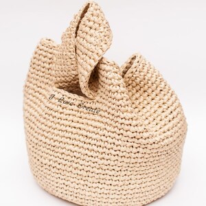 Pu&ograve; includere: Una borsa tote in rafia beige intrecciata con base arrotondata e parte superiore piegata. La borsa ha un aspetto strutturato e il testo "I Love Create" &egrave; visibile sul davanti.