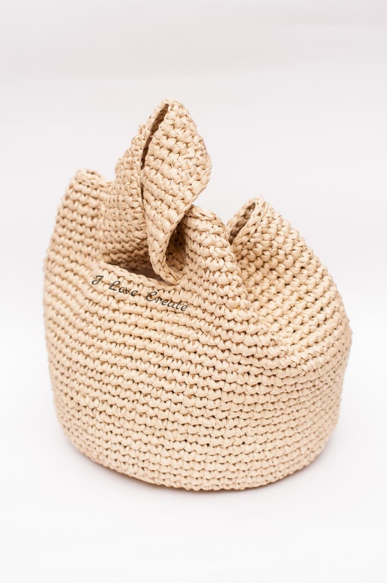 Pu&ograve; includere: Una borsa tote in rafia beige intrecciata con base arrotondata e parte superiore piegata. La borsa ha un aspetto strutturato e il testo "I Love Create" &egrave; visibile sul davanti.