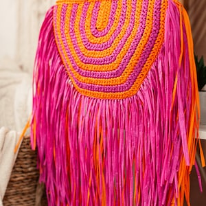 May include: A vibrant, handmade straw bag with a bamboo handle. The bag features a half-circle design with alternating orange and pink stripes. Long pink and orange fringe hangs from the bottom, creating a playful, bohemian look.