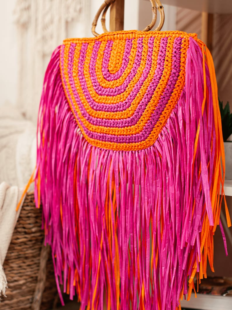 May include: A vibrant, handmade straw bag with a bamboo handle. The bag features a half-circle design with alternating orange and pink stripes. Long pink and orange fringe hangs from the bottom, creating a playful, bohemian look.