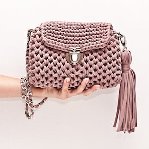 Puede incluir: Un bolso de mano de crochet color malva con una correa de cadena plateada y un borla. El bolso tiene un cierre de broche plateado.