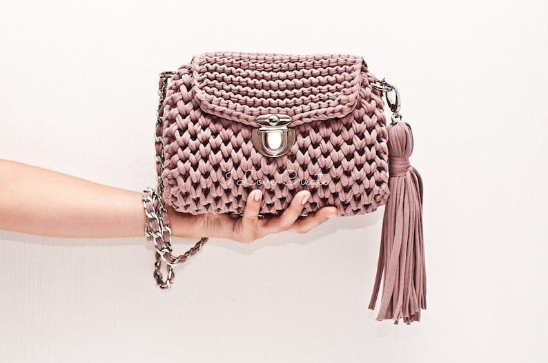 Puede incluir: Un bolso de mano de crochet color malva con una correa de cadena plateada y un borla. El bolso tiene un cierre de broche plateado.