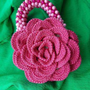 May include: A pink crocheted flower-shaped handbag with a beaded handle. The handle is made of pink and white beads.