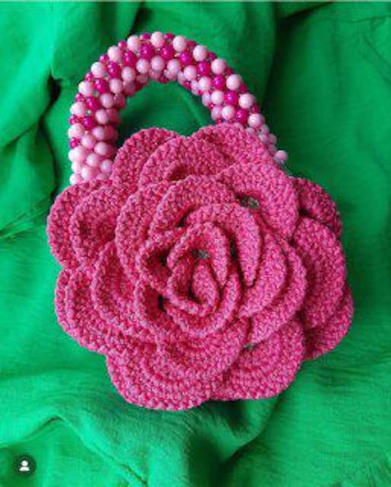 May include: A pink crocheted flower-shaped handbag with a beaded handle. The handle is made of pink and white beads.