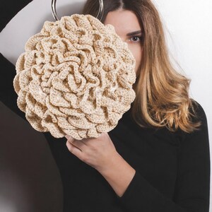 May include: A beige, round crocheted handbag with a silver metal handle. The bag has a textured, ruffled design. The person holding the bag is wearing a black long-sleeved shirt.