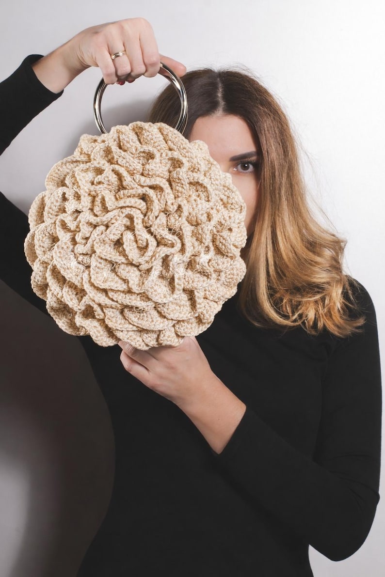 May include: A beige, round crocheted handbag with a silver metal handle. The bag has a textured, ruffled design. The person holding the bag is wearing a black long-sleeved shirt.