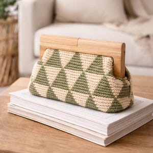 Op de afbeelding: Een handgemaakte gehaakte clutch met een geometrisch patroon in olijfgroen en cr&egrave;me. De tas heeft een houten frame en wordt getoond op een stapel boeken op een houten tafel. De achtergrond omvat een neutrale bank en een rieten mand.