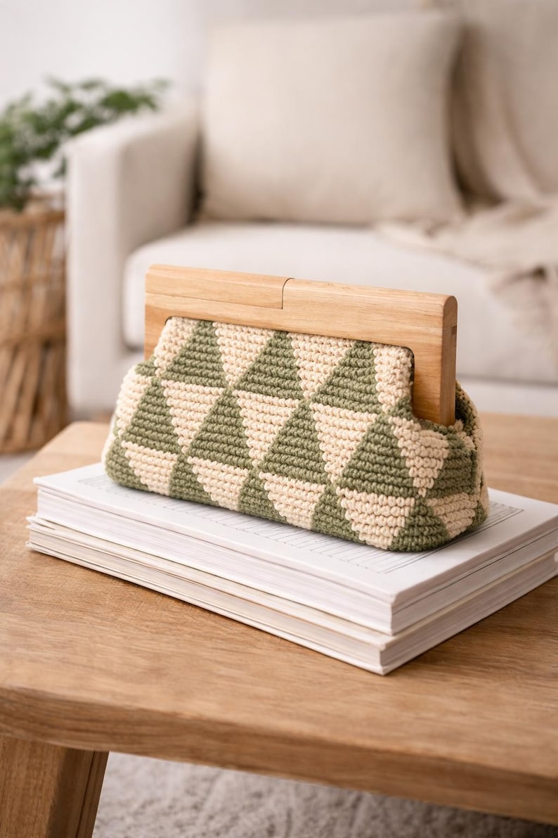 Op de afbeelding: Een handgemaakte gehaakte clutch met een geometrisch patroon in olijfgroen en cr&egrave;me. De tas heeft een houten frame en wordt getoond op een stapel boeken op een houten tafel. De achtergrond omvat een neutrale bank en een rieten mand.