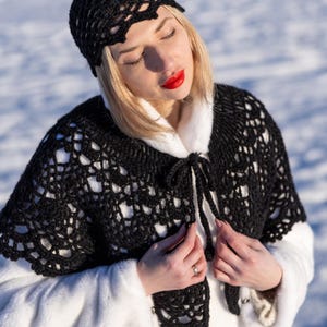 Könnte beinhalten: Ein schwarzer gehäkelter Schal und passender Hut. Das offene Design des Schals hat einen Krawattenverschluss vorne. Der Hut hat einen gewellten Rand. Der Schal wird über einem weißen Kunstfellmantel getragen.