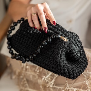 May include: A black crocheted clutch with a beaded strap. The clutch has a textured appearance and a gold-colored clasp. The strap features black beads of various shapes. The overall design is elegant and suitable for evening wear.