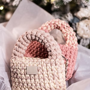 Op de afbeelding: Twee gehaakte handtassen in roze en cr&egrave;me met een zilveren kettingband. De kleinere tas heeft een label met de tekst "J'ois C'est"