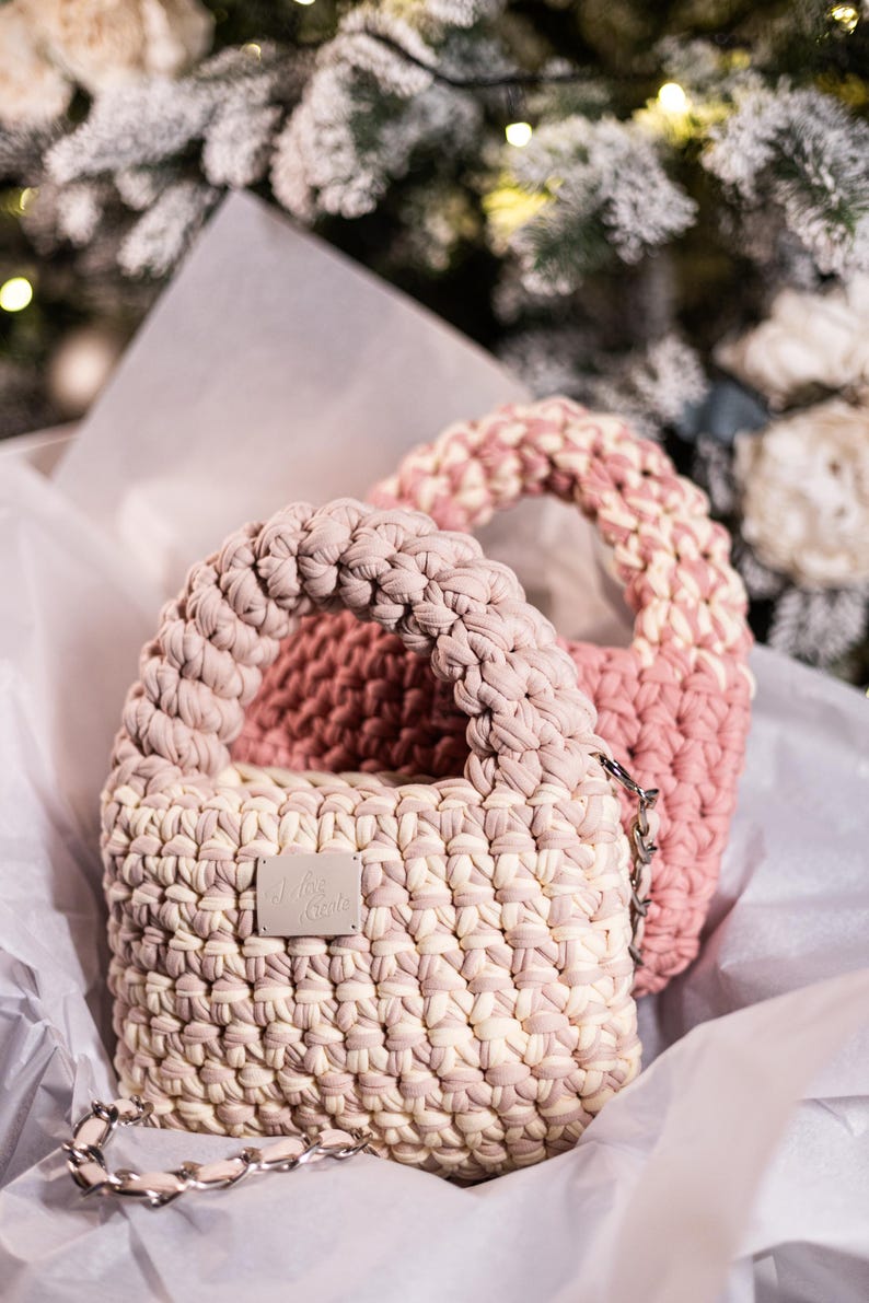 Op de afbeelding: Twee gehaakte handtassen in roze en cr&egrave;me met een zilveren kettingband. De kleinere tas heeft een label met de tekst "J'ois C'est"