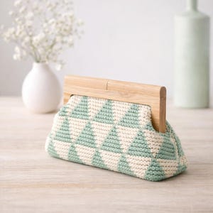 Op de afbeelding: Een gehaakte clutch met een geometrisch patroon in mintgroen en cr&egrave;me. De tas heeft een houten handvat en een driehoekige vorm. De clutch wordt gepresenteerd op een licht houten oppervlak.