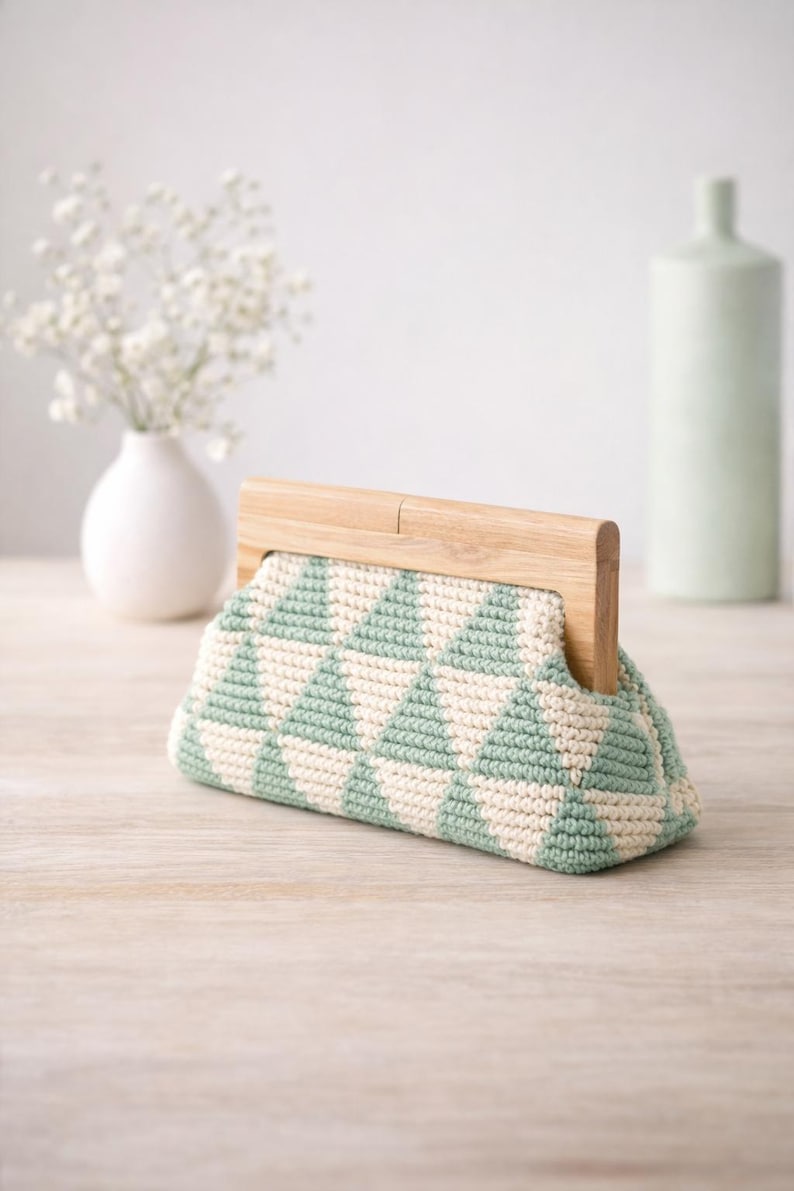 Op de afbeelding: Een gehaakte clutch met een geometrisch patroon in mintgroen en cr&egrave;me. De tas heeft een houten handvat en een driehoekige vorm. De clutch wordt gepresenteerd op een licht houten oppervlak.
