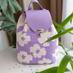 Puede incluir: Mochila de crochet hecha a mano con un estampado floral en lavanda y crema. La bolsa presenta una solapa y un asa de cuero lavanda, aseguradas con un cierre dorado. La bolsa está sobre una mesa blanca.