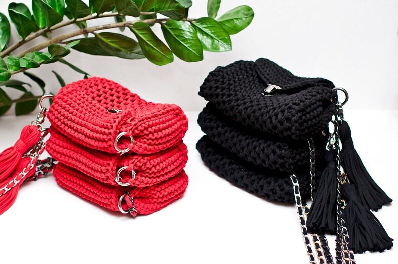 Puede incluir: Dos bolsos de crochet, uno rojo y otro negro, con correas de cadena y borlas. Los bolsos est&aacute;n apilados uno encima del otro.