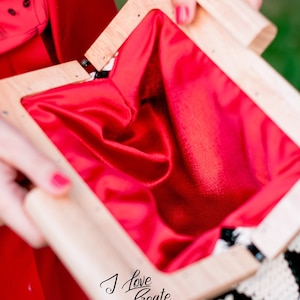 Op de afbeelding: Een handtas met houten frame en een felrode satijnen voering. De tas is open en toont de zachte binnenkant. Het frame is gemaakt van lichtgekleurd hout. De tekst "I Love Create" is zichtbaar.