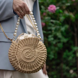 May include: A circular, beige crochet handbag with a spiral pattern. It has a pearl beaded handle and a drawstring closure. The bag is held by a person wearing a light blue blazer.