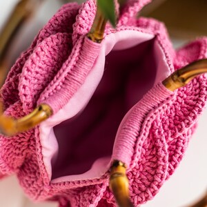 May include: A pink crocheted bag with bamboo handles. The bag has a ruffled design and a pink lining.