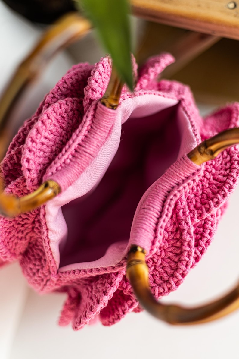 May include: A pink crocheted bag with bamboo handles. The bag has a ruffled design and a pink lining.