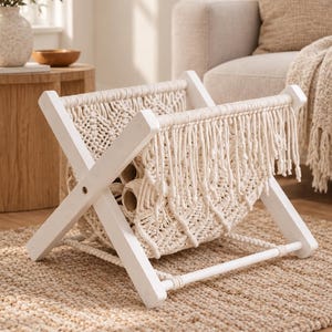 May include: A white wooden X-frame magazine rack featuring a cream-colored macrame sling with intricate knotted patterns and long fringe detailing hanging from the top bars, designed to hold items in a suspended fabric cradle.