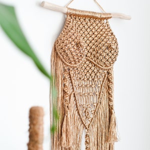 Könnte beinhalten: Ein goldener Makramee-Wandbehang mit einem einzigartigen Design und Fransen-Details. Der Wandbehang ist aus Naturfasern gefertigt und hat eine Boho-Ästhetik.