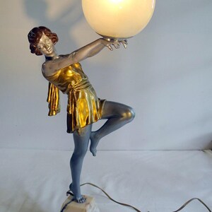 Peut inclure: Lampe de style Art Déco représentant une figure féminine stylisée en robe dorée tenant un grand globe lumineux. La figurine est argentée et bleue, sur un socle en marbre. La lampe a une esthétique vintage.