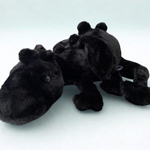 Peut inclure: Une peluche en forme de dragon noir avec une fourrure douce, une longue queue et de petites ailes sur le dos. La créature a un museau arrondi, quatre pattes avec des griffes visibles et de petits yeux verts contrastant avec le tissu sombre.