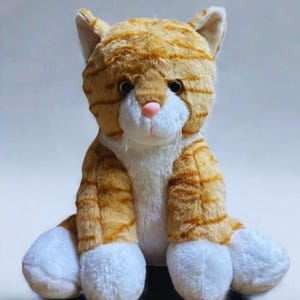 May include: A plush cat toy featuring orange and white striped fur. The stuffed animal has a white chest, white paws, and a small pink nose. It is depicted in a seated position with dark eyes and white whiskers, crafted from soft, textured fabric.