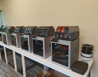 Könnte beinhalten: Fünf 3D-Drucker in einer Reihe auf einem weißen Tisch. Die Drucker sind silber und schwarz mit durchsichtigen Plastikdeckeln. Jeder Drucker hat eine Filamentspule oben. Die Drucker befinden sich in einem Raum mit weißen Wänden.