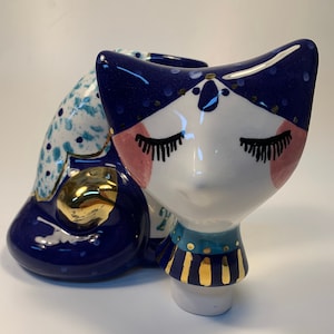 Könnte beinhalten: Eine Keramik-Katzenfigur, die in Blau und Weiß mit goldenen Akzenten bemalt ist. Die Katze liegt mit dem Kopf auf den Pfoten und hat die Augen geschlossen.