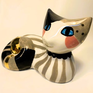 May include: A ceramic cat figurine with black, white, and gray stripes. The cat has blue eyes and pink cheeks. The cat is lying down with its head turned to the side. The cat has a gold ring around its neck.