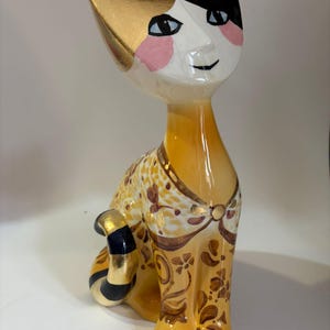 May include: A ceramic cat figurine with a white face, black ears, and a gold-painted body. The cat is wearing a yellow and brown patterned dress with gold accents. The cat is sitting with its tail curled around its legs.